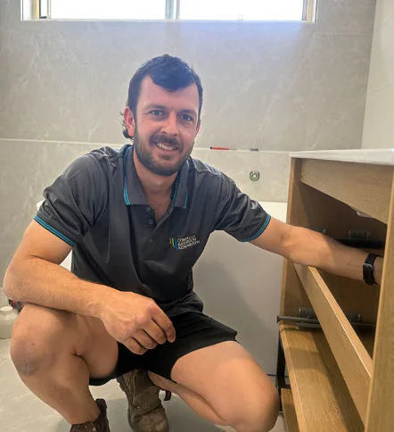 Smiling bathroom renovation worker in branded uniform crouching beside partially installed wooden vanity unit in a tiled bathroom.