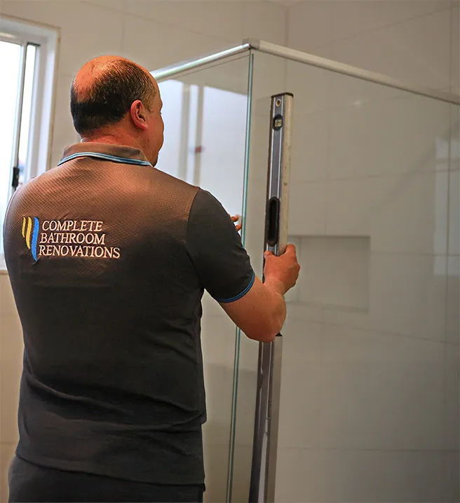Bathroom renovation specialist from Complete Bathroom Renovations aligning a glass shower screen using a spirit level during installation inside a newly tiled modern bathroom.