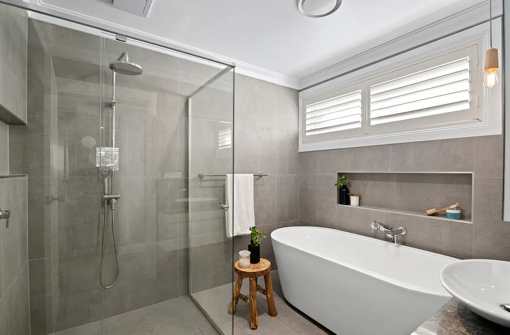 Contemporary bathroom makeover with freestanding bathtub, walk-in shower with glass screen, grey porcelain tiles, recessed wall niche, white plantation shutters, and minimalist timber stool styling.