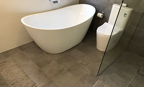 Freestanding bath surrounded by soft grey tiles and chrome fixtures under natural light.