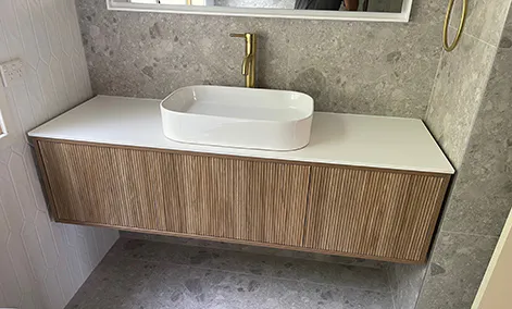 Timber vanity with white countertop, round basin, and brushed gold tapware against light tiled walls.
