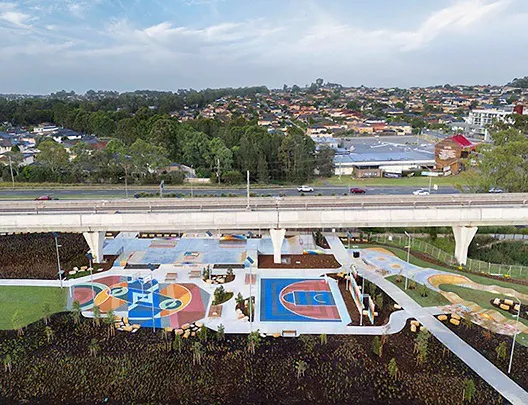Aerial view of Beaumont Hills parklands featuring colourful basketball courts, walking paths, and landscaped green spaces beneath the metro line bridge, with suburban housing in the background.