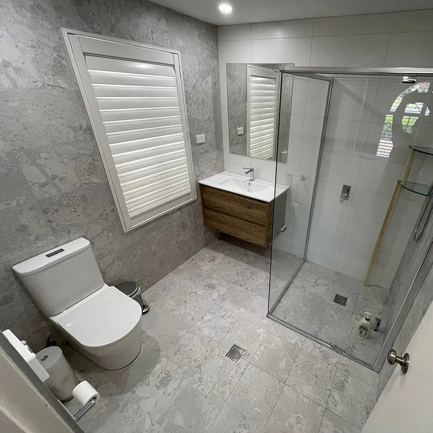Contemporary bathroom renovation in Beaumont Hills with grey stone-look tiles, floating timber vanity, glass shower enclosure, and white plantation shutters for a clean, modern aesthetic.