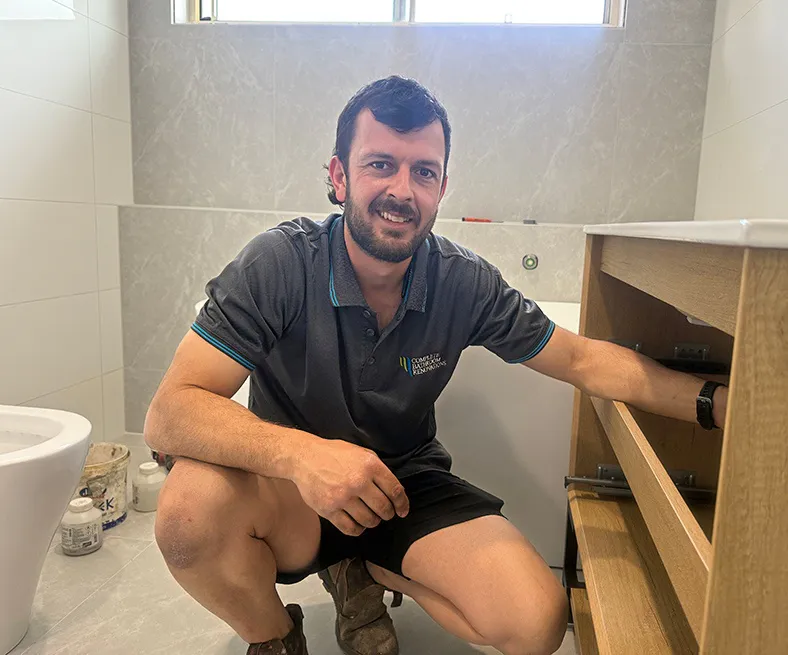 Bathroom renovation specialist kneeling beside a timber vanity unit during installation, wearing a branded Complete Bathroom Renovations shirt, with light grey tiled walls and floor visible in the background.