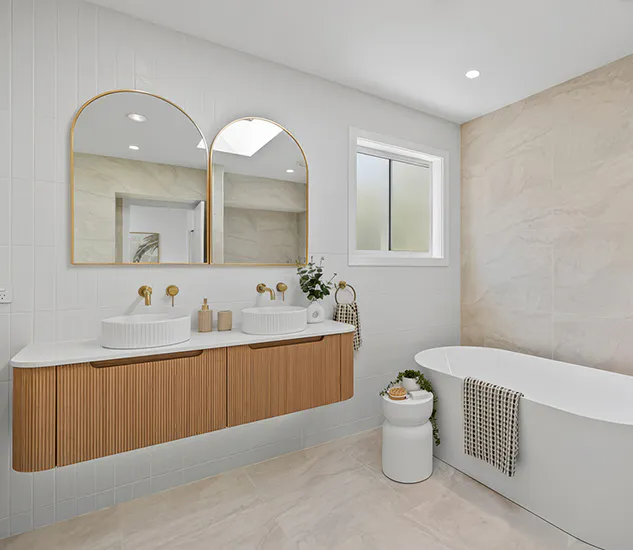 Bright contemporary bathroom with fluted timber double vanity, gold wall-mounted tapware, twin arched mirrors, and a white freestanding bath against soft beige tiles, styled for a clean modern look.