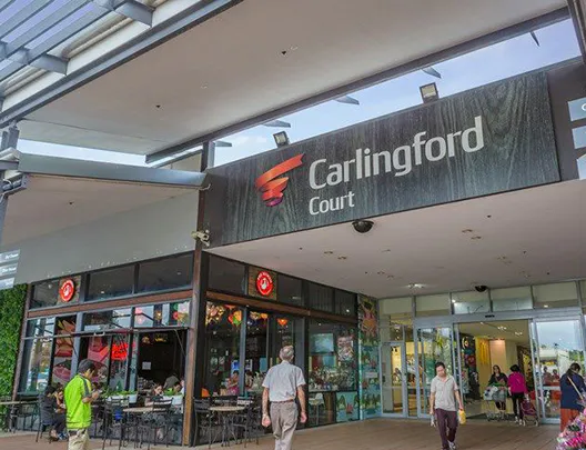 Street-level view of Carlingford Court shopping centre entrance with shoppers walking by and café seating in front.