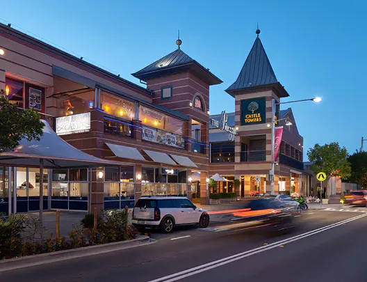Evening street view of Castle Towers in Castle Hill, showing illuminated shopfronts, passing traffic, and the building’s distinctive peaked rooftops under a clear twilight sky.