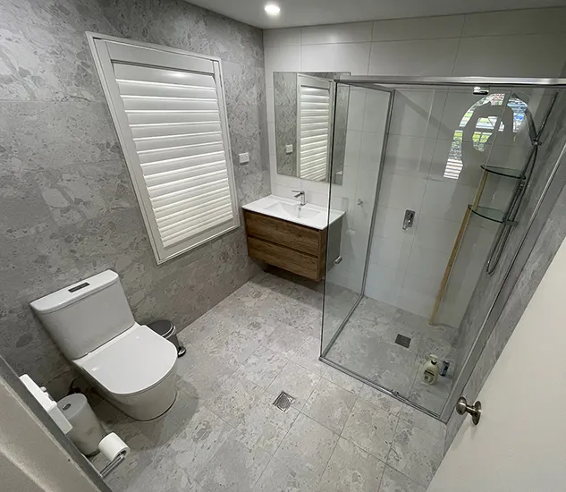 Compact modern bathroom featuring a glass-enclosed shower, wall-hung timber vanity, large mirror, and stone-look tiles for a bright, contemporary feel.