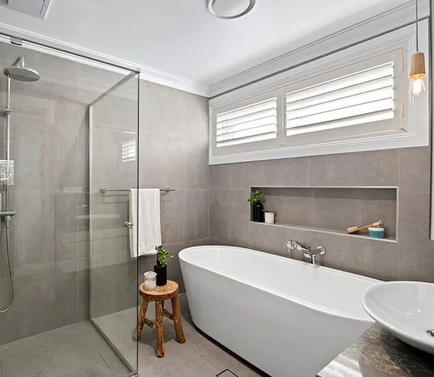 Contemporary bathroom featuring a freestanding white bathtub, walk-in glass shower with rainfall head, grey floor-to-ceiling tiles, recessed wall shelf, plantation shutters, and warm timber accents creating a calm, modern atmosphere.
