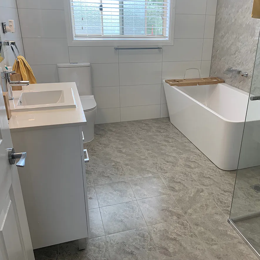 Contemporary bathroom with a sleek white freestanding bath, glass shower screen, white vanity with chrome tapware, grey marble-look floor tiles, and neutral wall tiles complemented by natural light from a window with blinds.