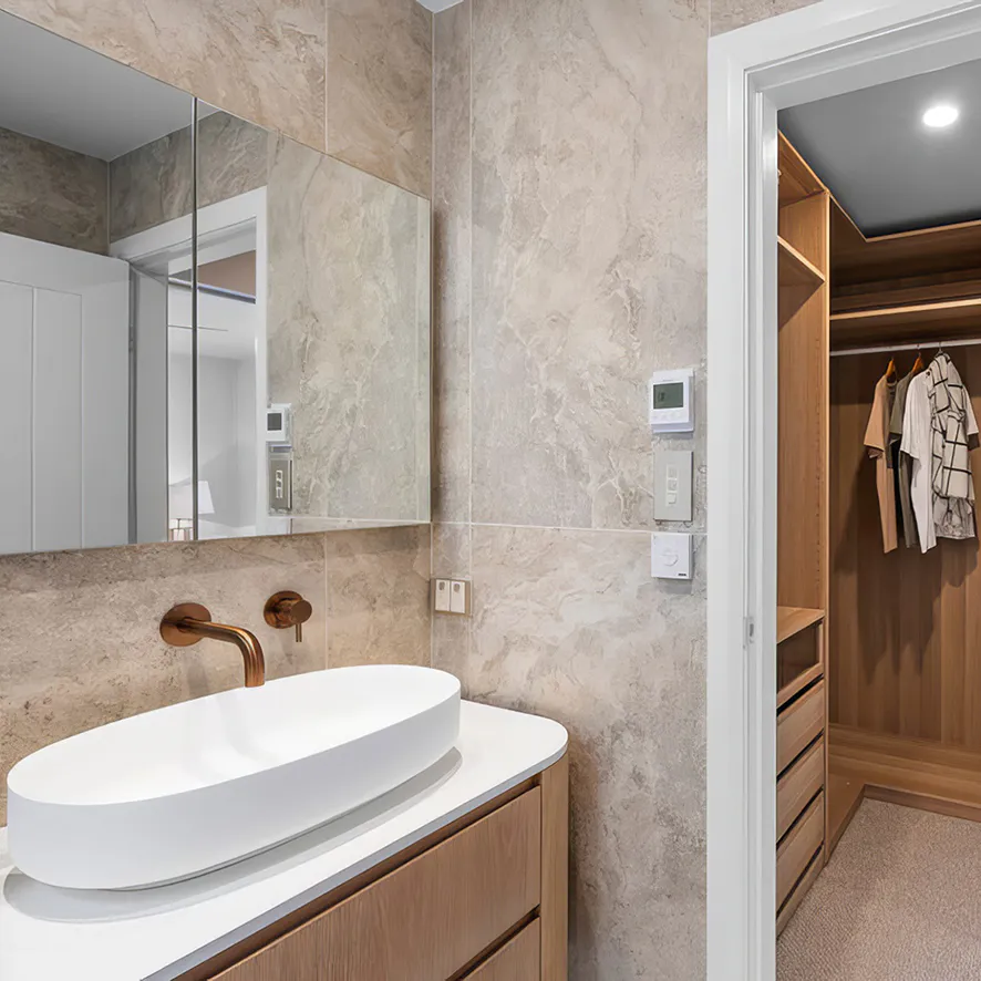 Modern ensuite featuring a wall-mounted vanity with a white vessel basin and brushed bronze tapware, opening into a custom timber walk-in wardrobe with built-in shelving and soft lighting.