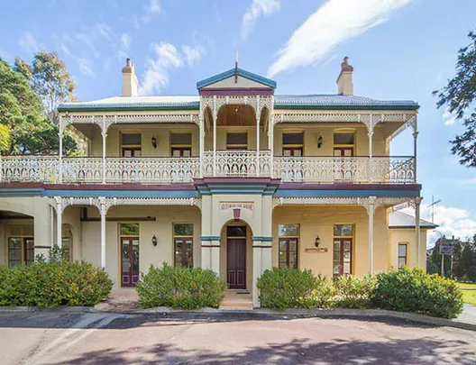 Exterior of Glenwood House in New South Wales, a two-storey heritage building with ornate wrought-iron balconies, cream facade, green trim, and manicured gardens under a bright blue sky.