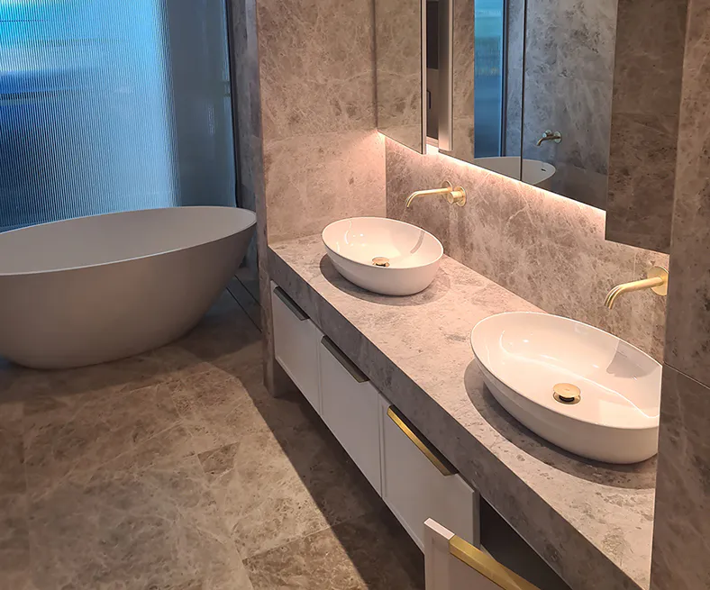 Luxury bathroom featuring double vanity with marble benchtop, twin vessel basins, brushed brass tapware, soft backlighting, and a freestanding bathtub set against textured stone tiles.