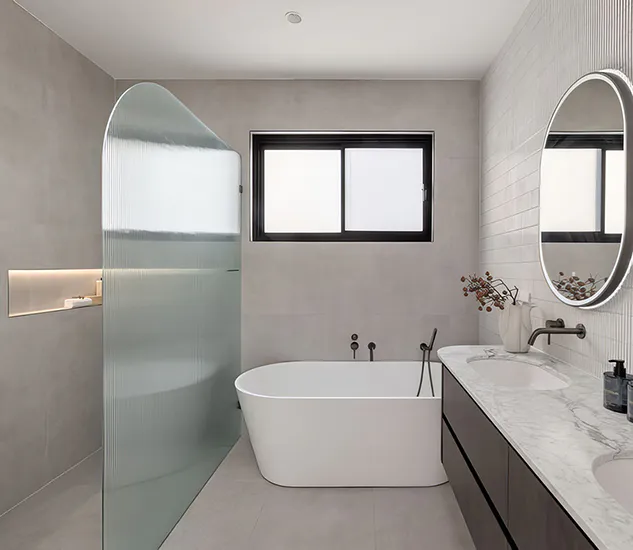 Luxury bathroom featuring a freestanding white bathtub beside a curved fluted glass shower screen, marble double vanity with LED backlit mirrors, matte black tapware, and soft grey wall tiles creating a refined, spa-like feel.