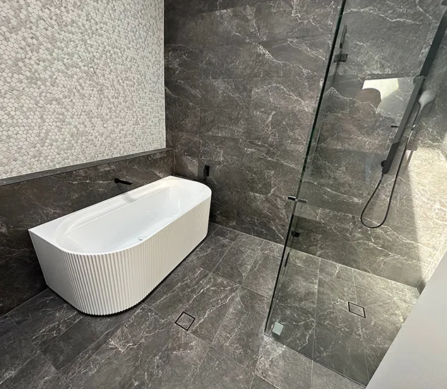 Luxury bathroom featuring a fluted freestanding white bath, dark marble-look floor and wall tiles, frameless glass shower with matte black fittings, and a mosaic feature wall adding texture and contrast.