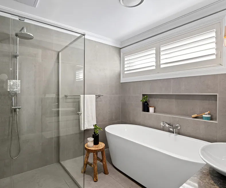 Modern bathroom in Beecroft featuring a freestanding bathtub, frameless glass shower, grey porcelain tiles, built-in wall niche, and plantation shutters for a calm, contemporary style.