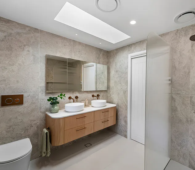 Contemporary bathroom renovation in Beecroft featuring bronze tapware, floating timber vanity with twin basins, fluted glass shower screen, and soft stone-look wall tiles illuminated by a skylight.
