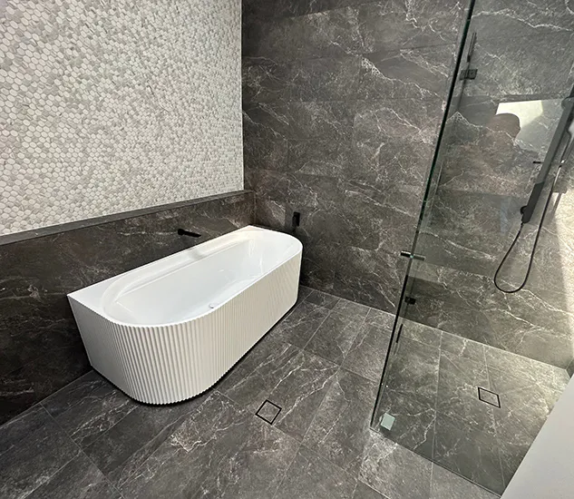 Contemporary bathroom renovation featuring a white fluted freestanding bath against dark marble wall and floor tiles, glass shower enclosure with black fixtures, and contrasting light mosaic feature wall.