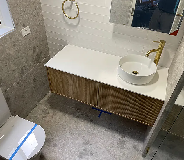 Modern bathroom with a floating timber vanity, white countertop and round vessel basin, brushed brass tapware and towel ring, grey stone-look tiles, and white subway tile splashback for a balanced, elegant finish.