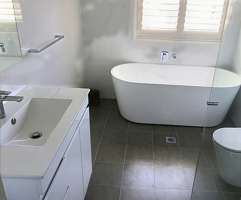 Modern bathroom with a white freestanding bathtub beneath plantation shutters, sleek vanity with integrated basin, frameless glass shower enclosure, and grey floor tiles creating a bright, minimalist space.