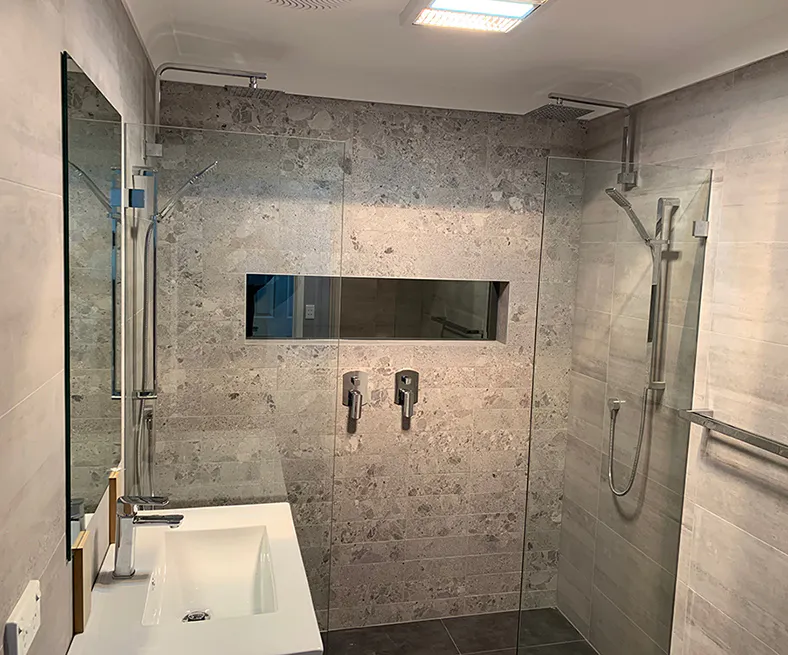Contemporary bathroom design featuring a frameless glass shower with dual rainfall and handheld shower heads, textured stone-look wall tiles, recessed shower niche, and sleek white vanity with chrome fixtures.