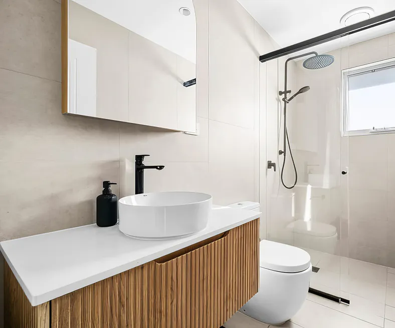 Modern bathroom featuring a frameless glass walk-in shower with matte black rainfall and handheld shower, wall-mounted timber vanity with white countertop basin, light beige wall tiles, and minimalist design with natural light from a window.
