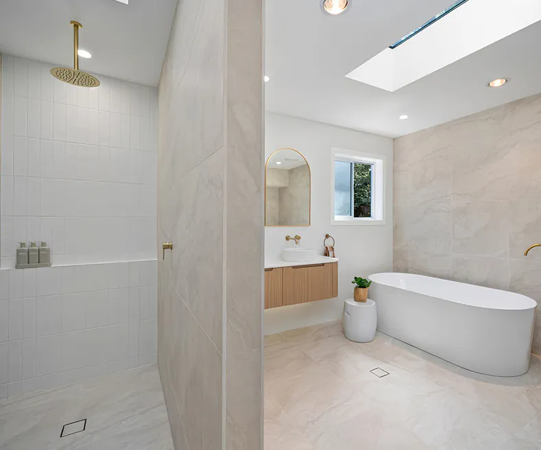 Luxurious Carlingford bathroom with freestanding bathtub, walk-in shower, skylight, gold tapware, and floating timber vanity against large neutral tiles.