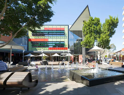 View of Rouse Hill Town Centre featuring the main fountain plaza with seating areas, shaded umbrellas, mature trees, and the colourful modern facade of the retail and dining precinct in the background.