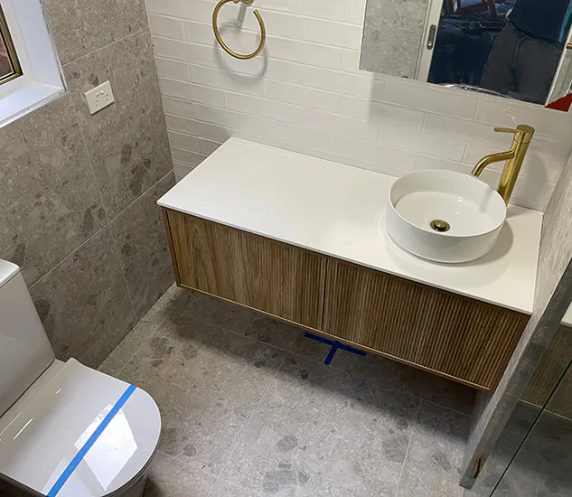 Compact Carlingford bathroom with a floating timber vanity, round countertop basin, brushed gold tapware, and grey tiled walls.