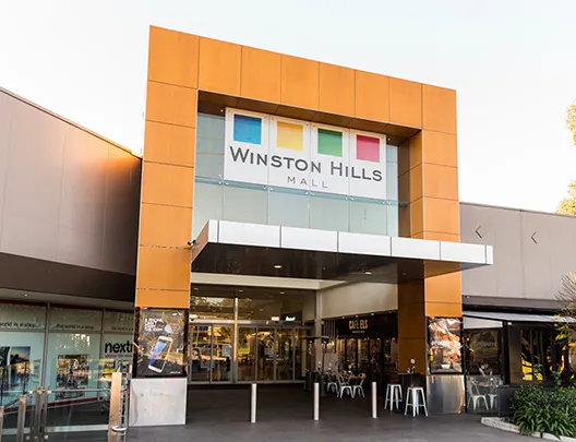 Exterior view of Winston Hills Mall shopping centre showing the main entrance with bold orange framing, glass panels, and signage above, with café seating and retail stores visible around the entry.