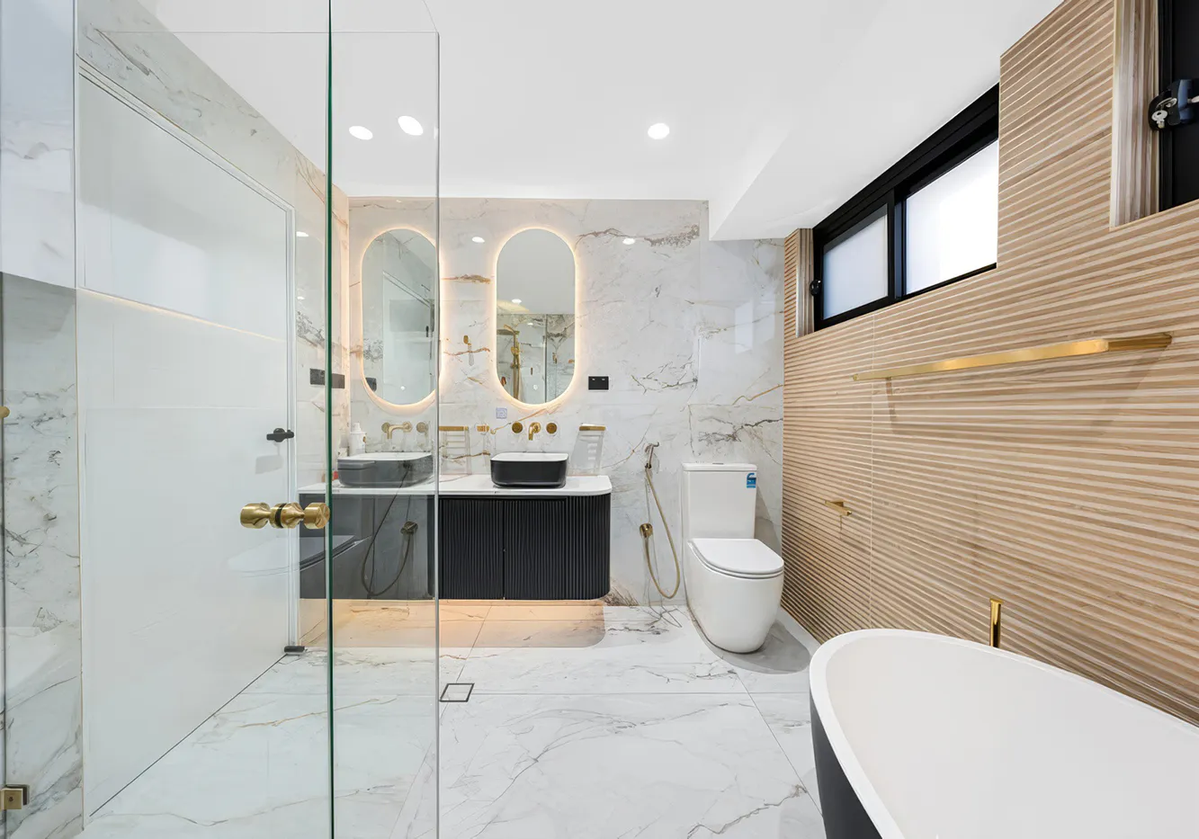 Luxurious bathroom with marble walls, modern freestanding bath, gold fixtures, and twin mirrors softly backlit — featured as the hero image for Complete Bathroom Renovations Carlingford.