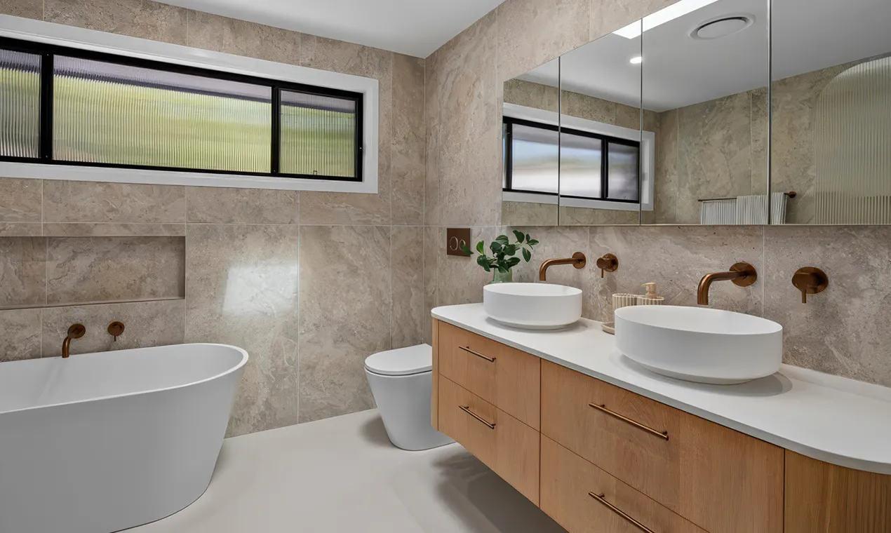 Luxury bathroom renovation in Winston Hills featuring a freestanding white bath, floating timber vanity with twin vessel basins, brushed brass tapware, large mirrored cabinets, and beige stone-look wall tiles creating a warm, contemporary aesthetic.