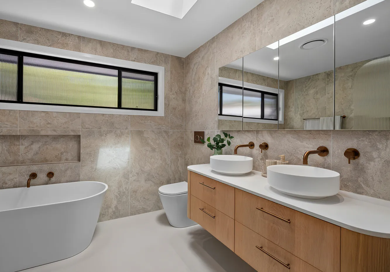 Luxury bathroom renovation in Winston Hills featuring a freestanding white bath, floating timber vanity with twin vessel basins, brushed brass tapware, large mirrored cabinets, and beige stone-look wall tiles creating a warm, contemporary aesthetic.