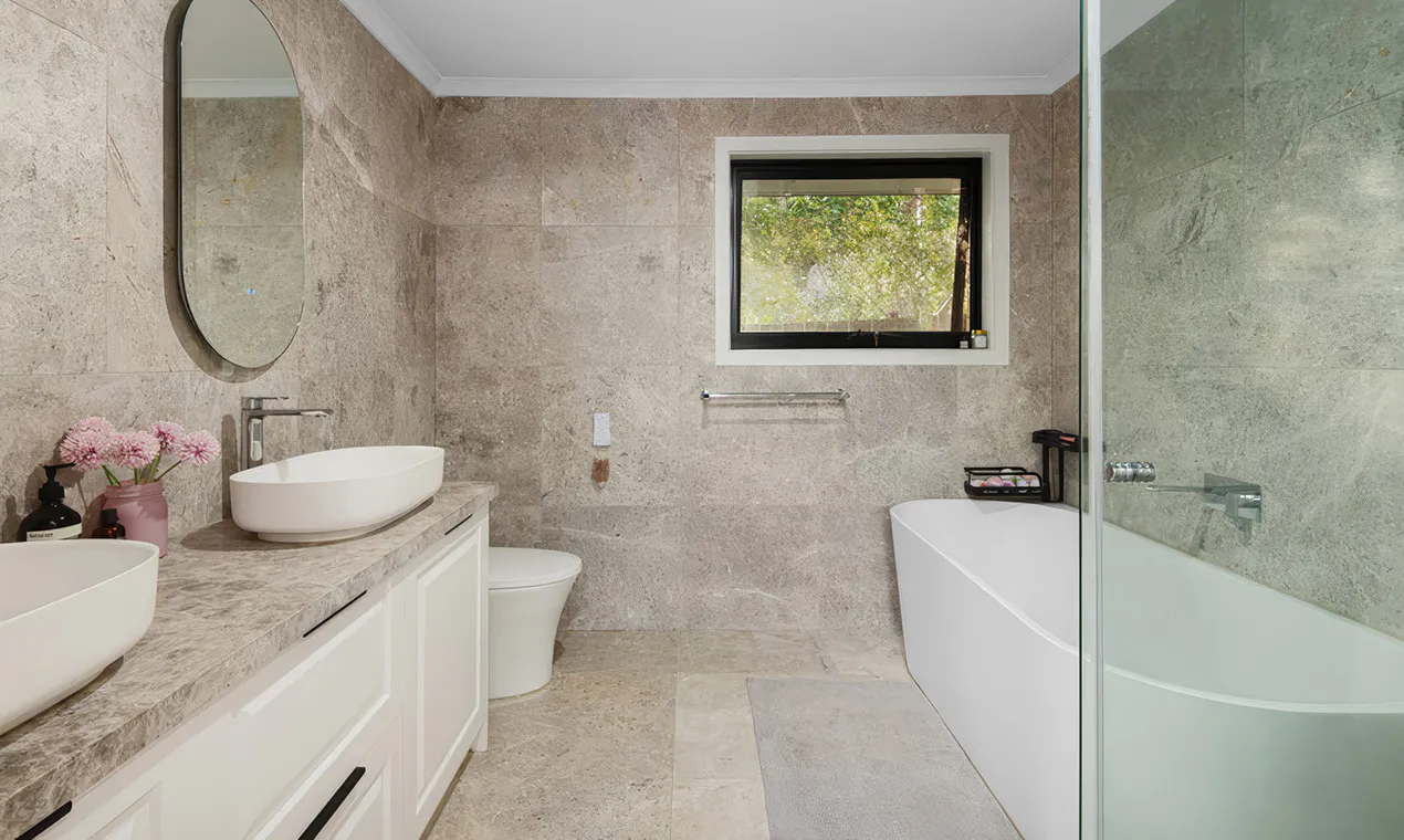Contemporary bathroom renovation in Castle Hill featuring a floating double vanity with white cabinetry, sleek chrome taps, large wall mirror, and grey stone-look tiles across the walls and floor, illuminated by a ceiling skylight.