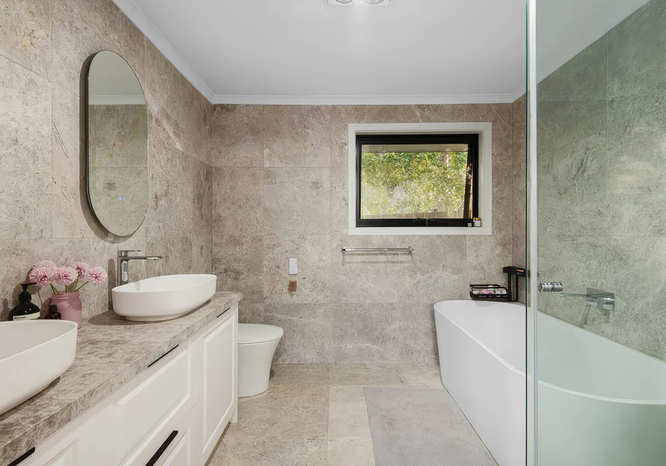 Contemporary bathroom renovation in Castle Hill featuring a floating double vanity with white cabinetry, sleek chrome taps, large wall mirror, and grey stone-look tiles across the walls and floor, illuminated by a ceiling skylight.
