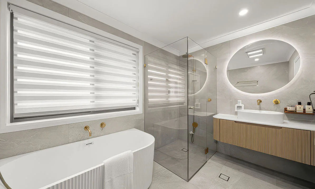 Modern bathroom with freestanding bathtub, glass shower, floating vanity with round backlit mirror, gold fixtures, and neutral stone tiles, used as a 'contact us' concept hero image.