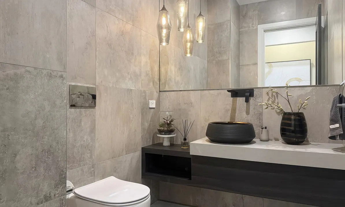 Modern bathroom renovation featuring large grey stone tiles, floating dark vanity with white countertop, black vessel basin, matte black tapware, and elegant pendant lighting creating a warm ambient glow.